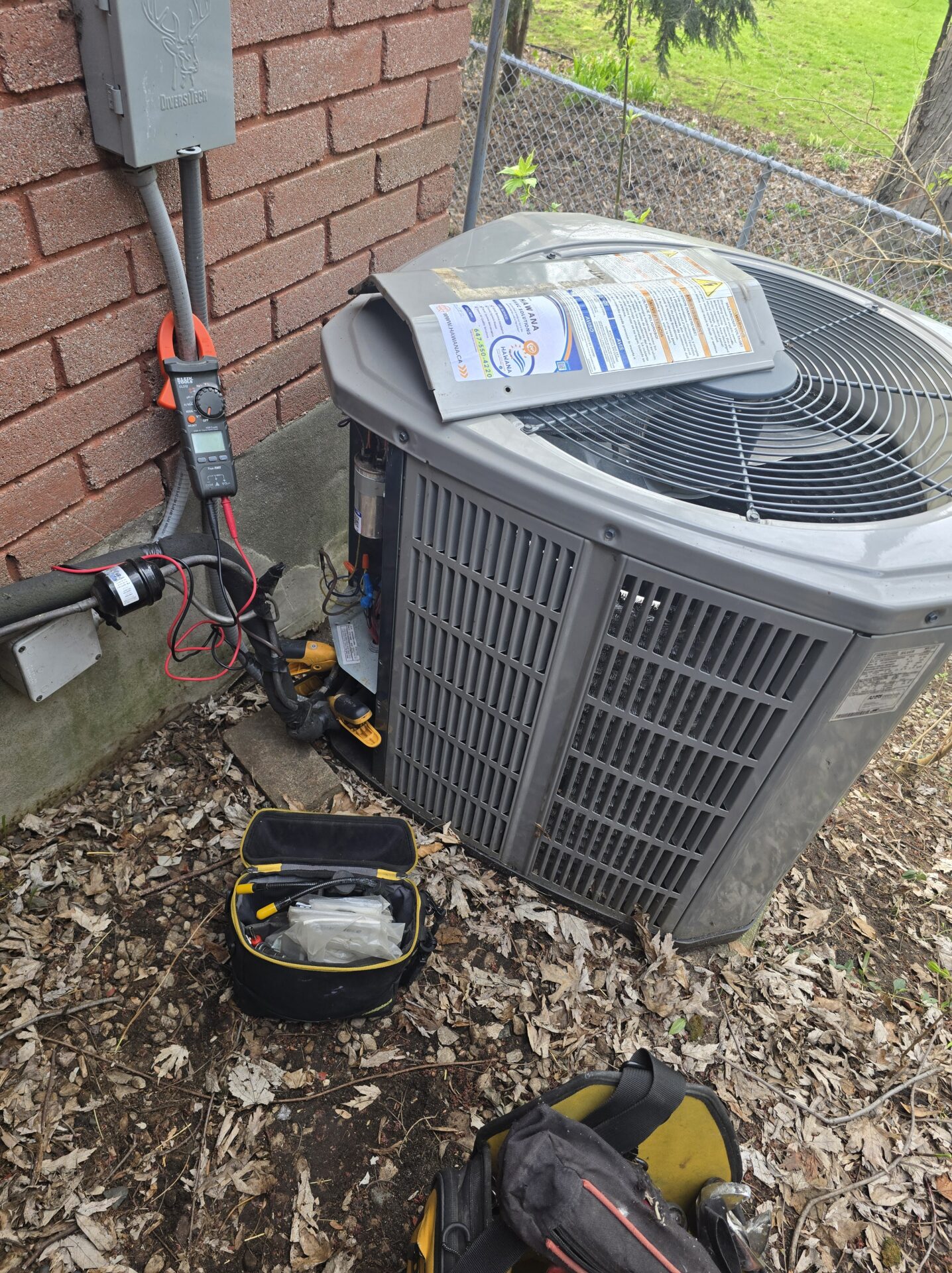 An outdoor air conditioning unit with the top panel removed, showing a Hawana HVAC Solutions service sticker, with a digital multimeter connected to the electrical components for testing.