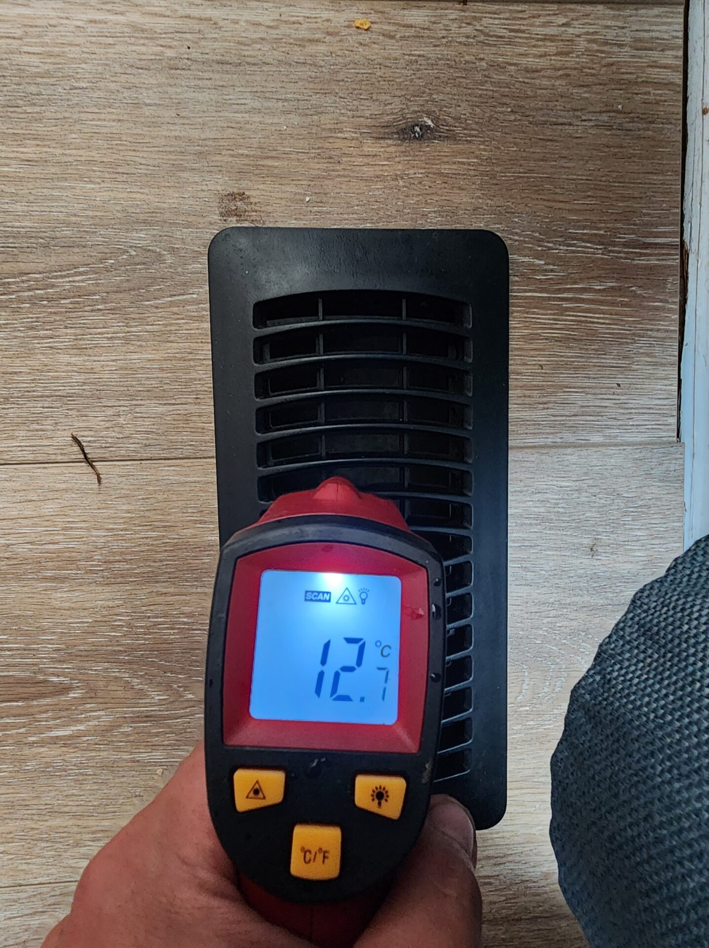 An infrared digital thermometer reading 12.7 degrees Celsius directly over a residential floor air vent.
