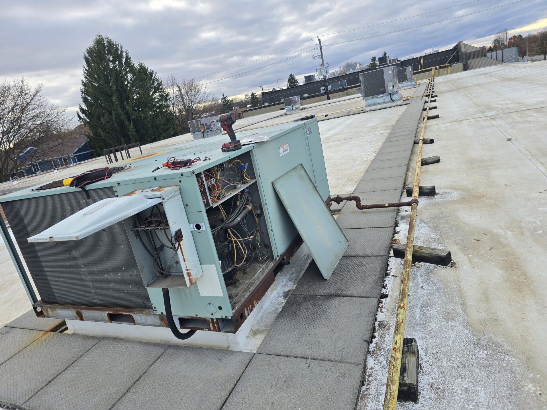 A large commercial rooftop HVAC unit with its side panels removed, revealing complex internal wiring, contactors, and the compressor compartment on a flat roof.