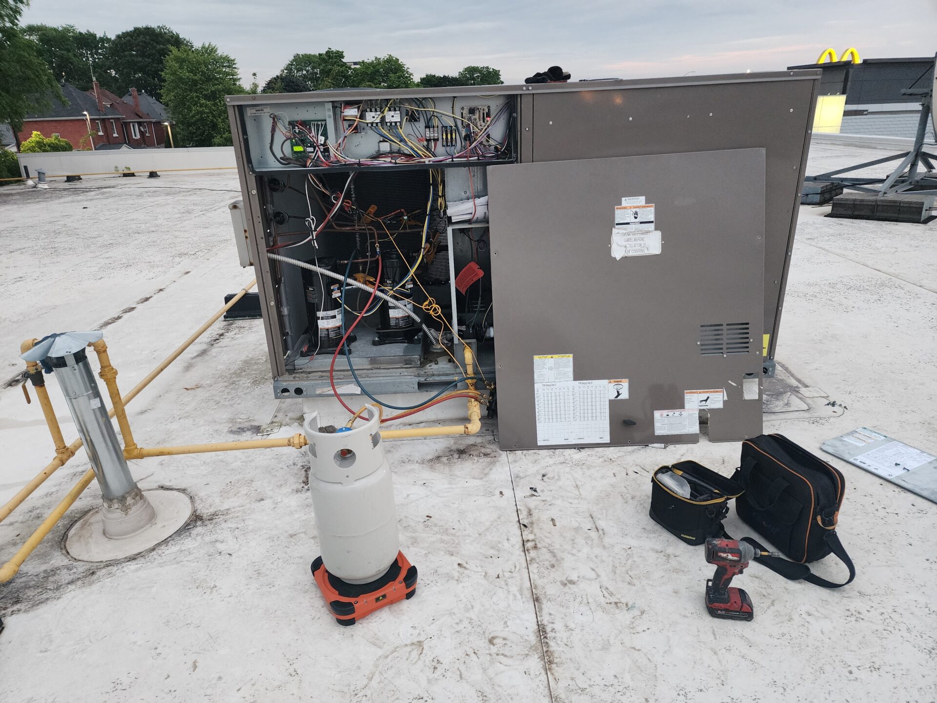 An open commercial rooftop HVAC unit (RTU) being serviced with a refrigerant recovery cylinder and manifold gauges attached