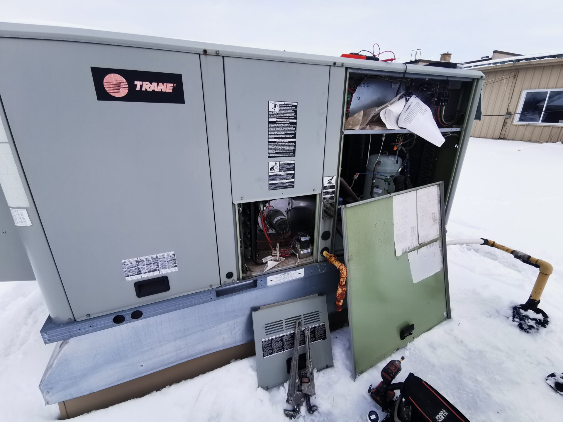 Open access panels on a Trane commercial rooftop HVAC unit being serviced during winter snow conditions.