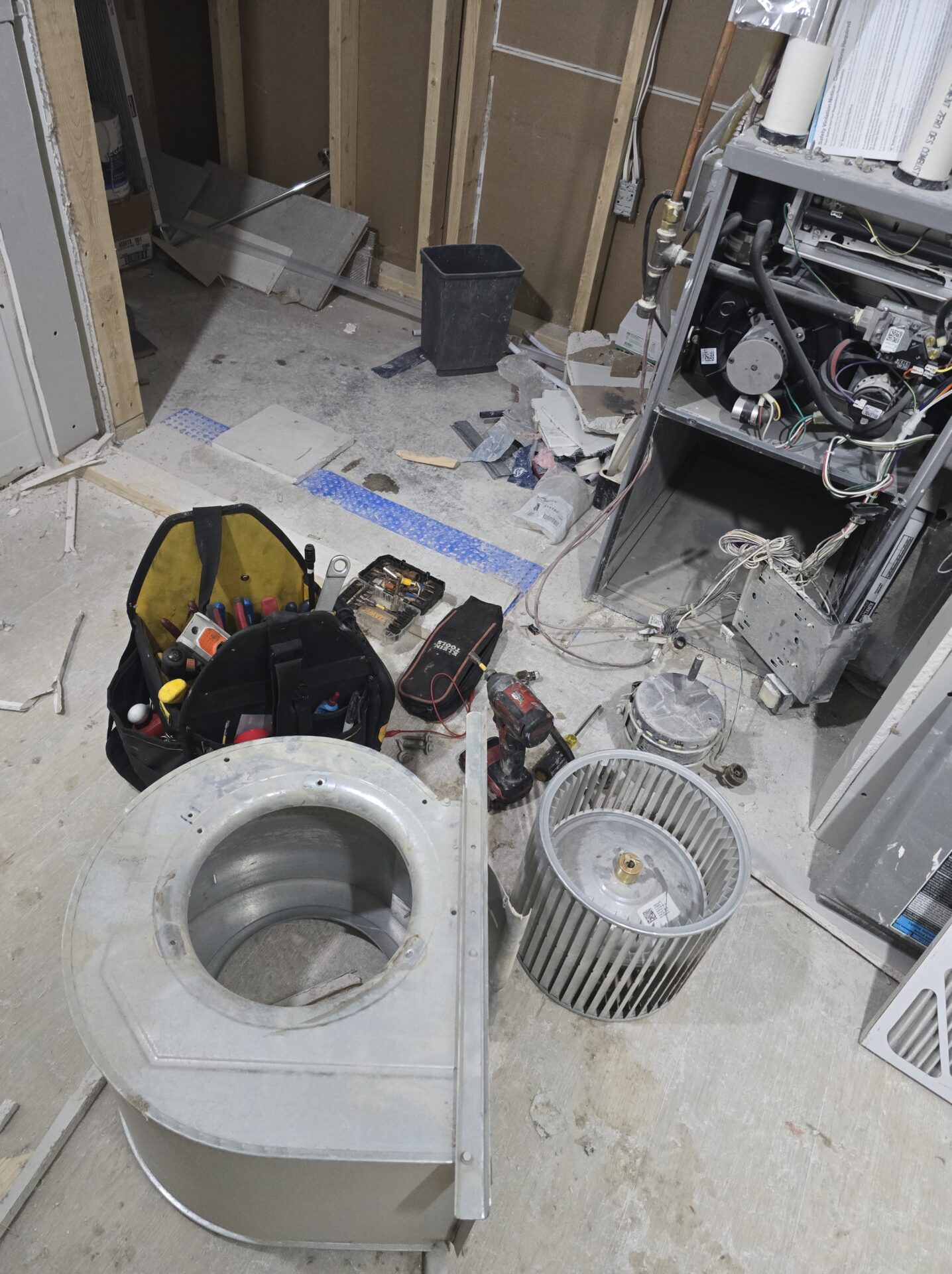 An open furnace in a basement with its blower wheel and motor fully removed and disassembled on the floor next to a professional HVAC tool bag.