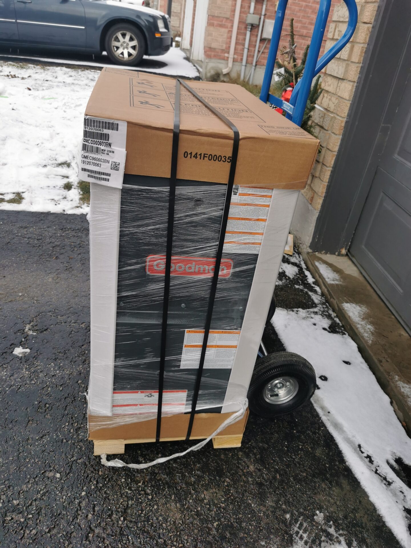 A brand new high-efficiency Goodman gas furnace packaged in a box and shrink-wrapped on a hand truck over a snow-dusted driveway.