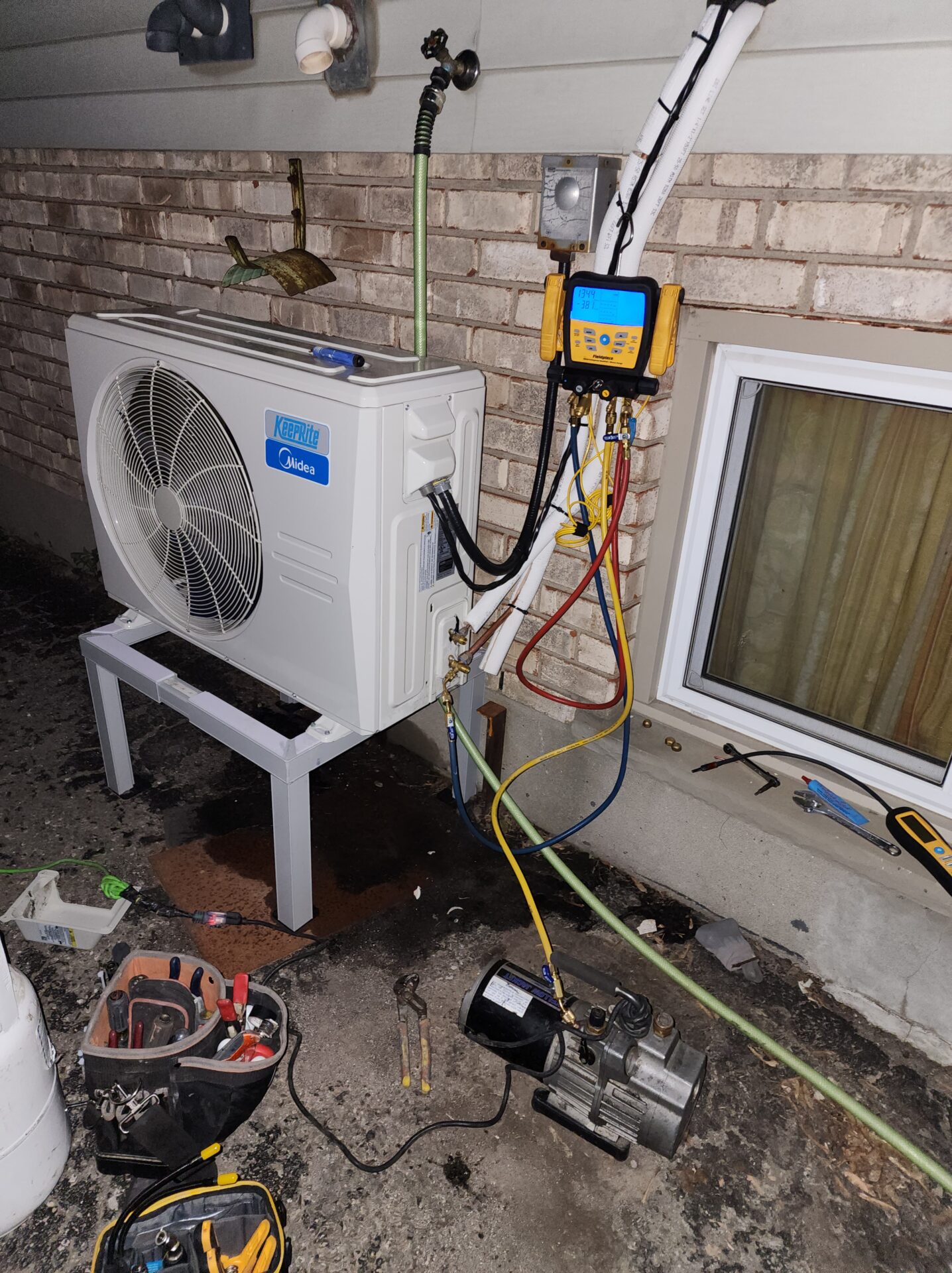 A KeepRite side-discharge heat pump unit on a snow stand undergoing deep system evacuation with a vacuum pump and digital gauges at night.