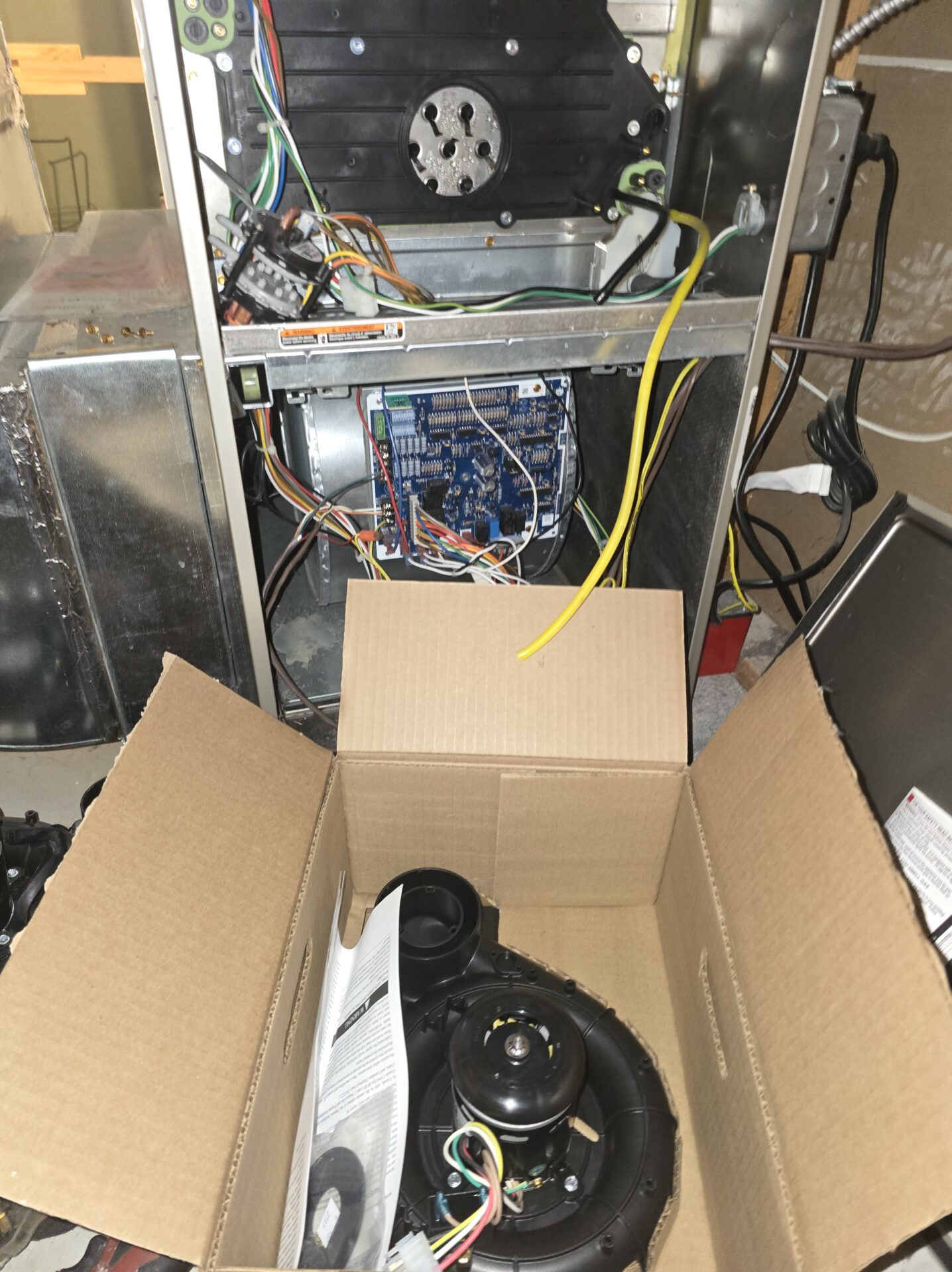 A brand new OEM furnace inducer blower motor assembly sitting in an open cardboard box, ready to be installed into the open gas furnace in the background.