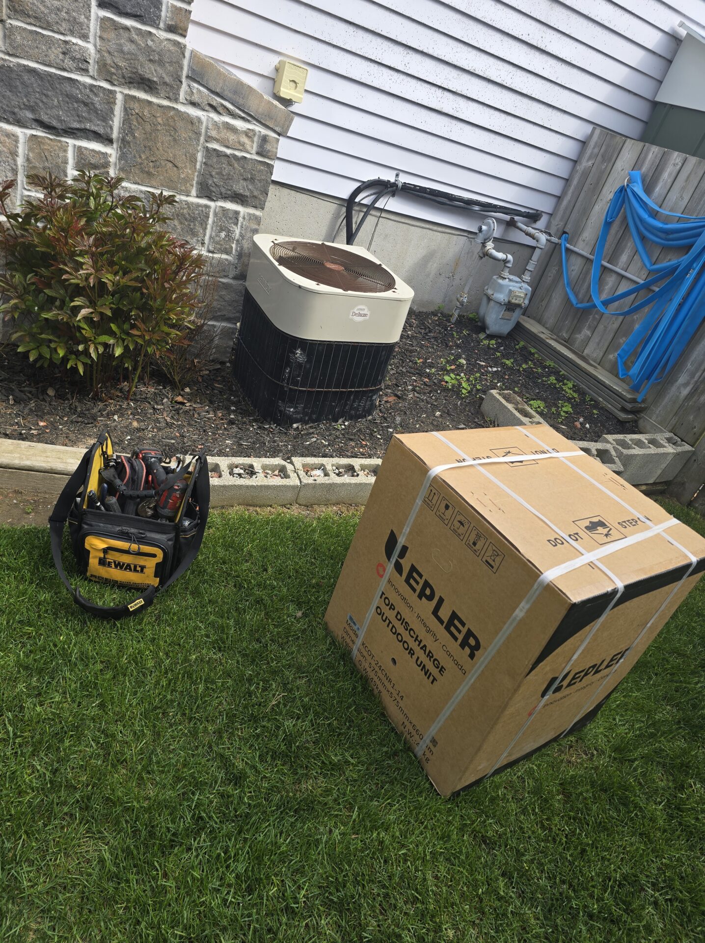 A brand new Kepler top discharge air conditioning unit in its shipping box sitting on a lawn next to an old AC unit and a technician's tool bag