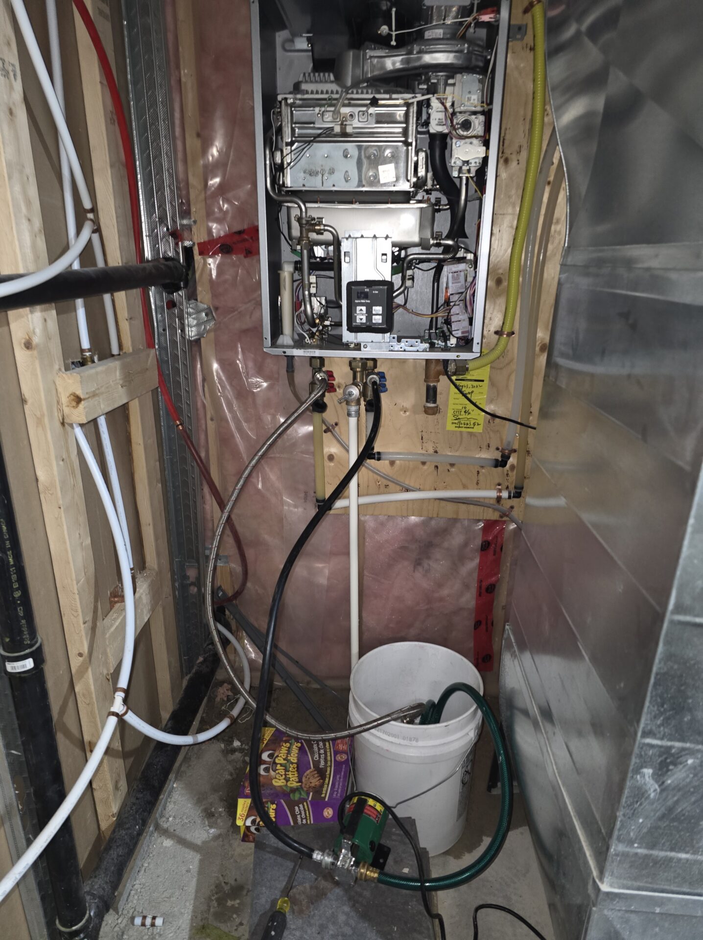 A tankless water heater undergoing descaling maintenance with a submersible pump, bucket, and hoses connected to the unit's isolation valves.