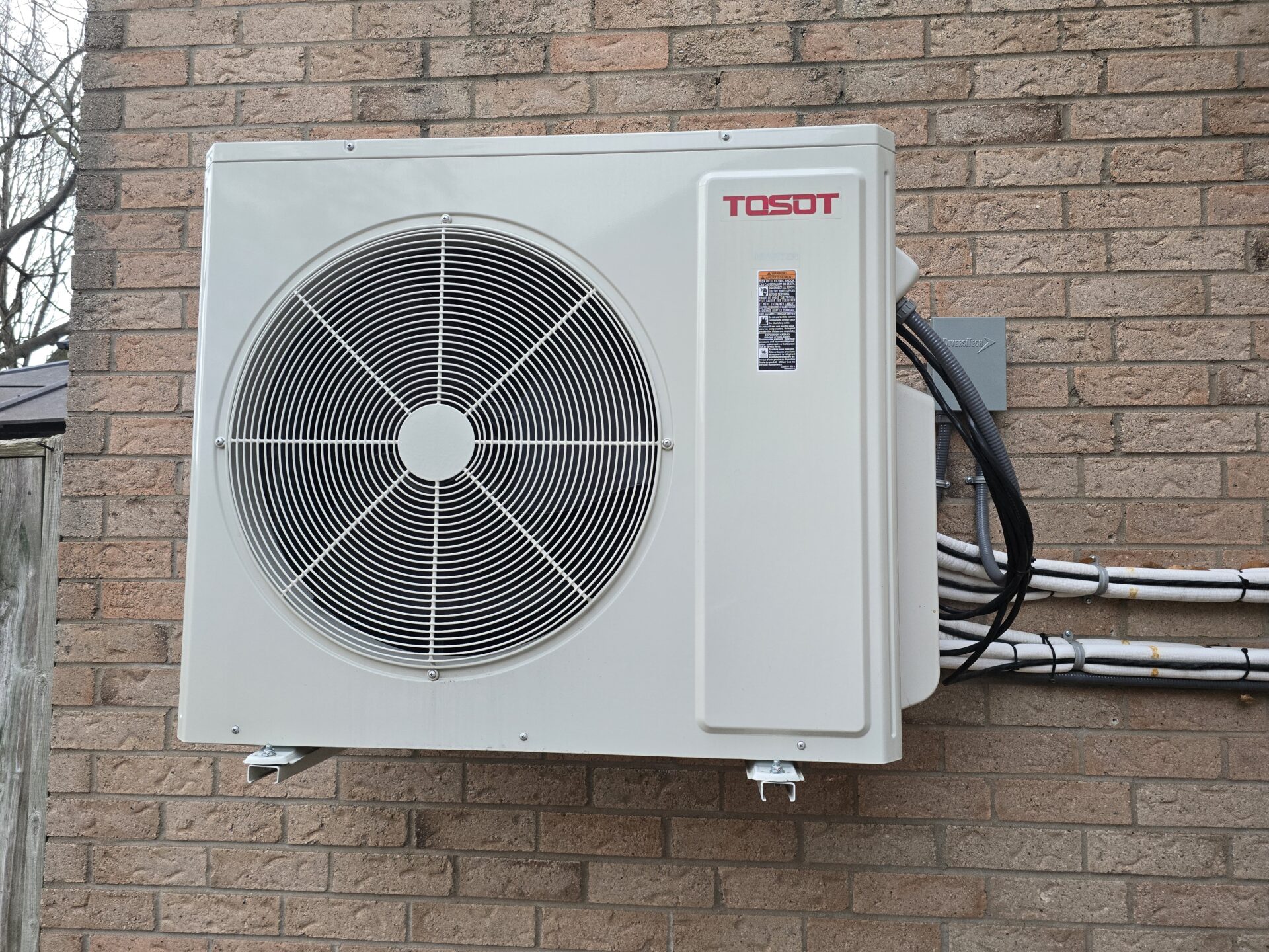 A new, white Multi zone TOSOT ductless mini-split outdoor condenser unit securely mounted on white brackets against a residential brick wall, with neatly organized, insulated refrigerant lines.