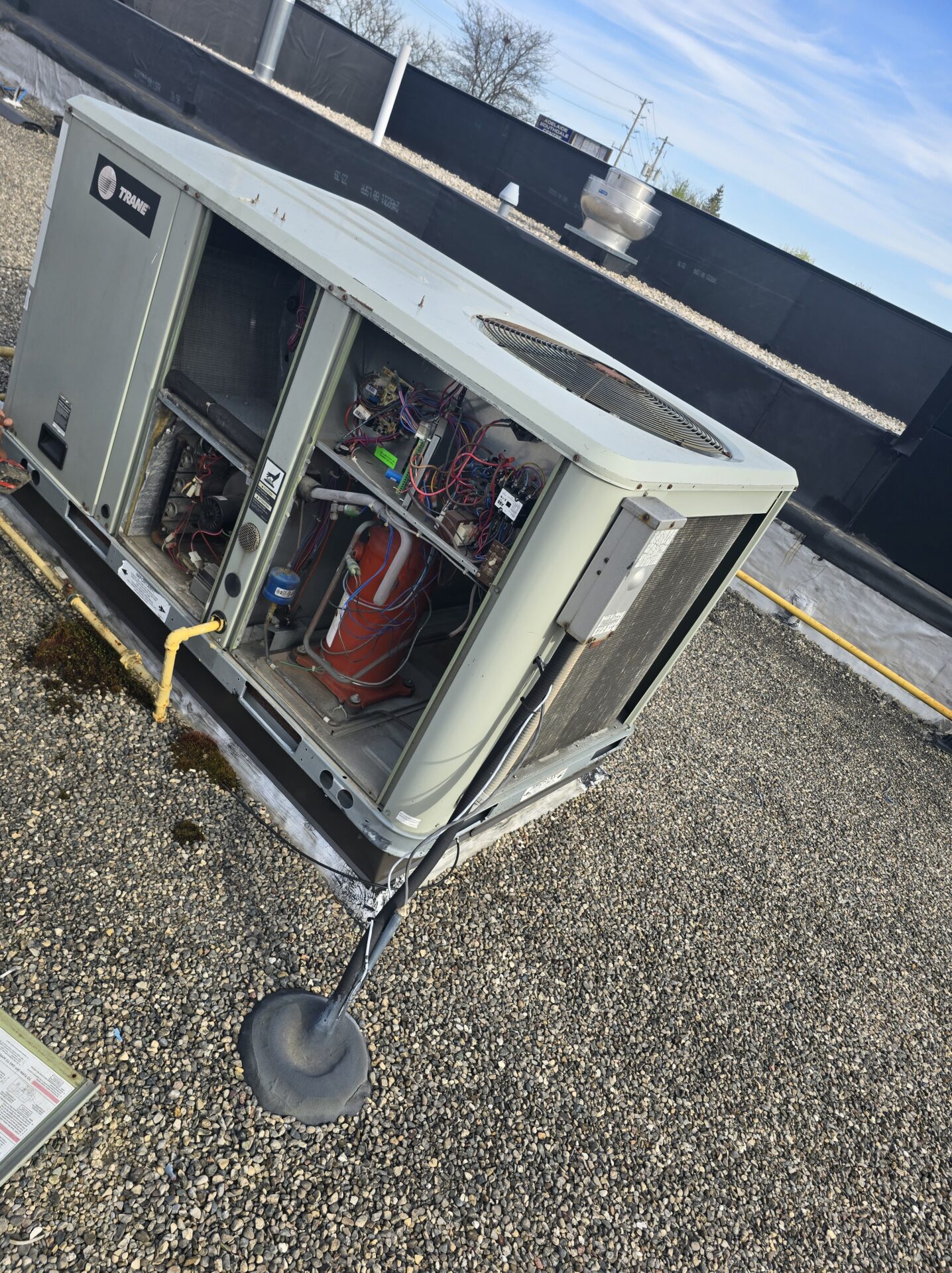An open Trane commercial rooftop HVAC unit sitting on a flat gravel roof, exposing internal wiring and the red compressor.