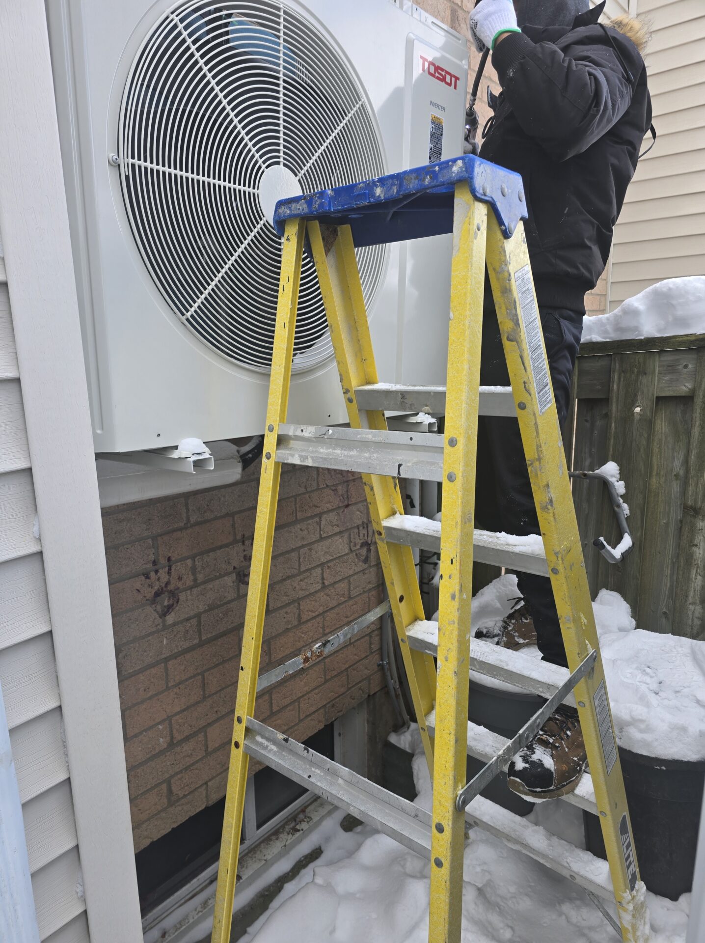 A technician standing on a yellow fiberglass step ladder in the snow, working on a wall-mounted TOSOT outdoor heat pump unit.